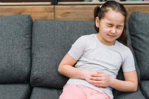 Menina no sofá com as mão no abdomen por estar sentindo uma dor abdominal e a cara com feições de quem está com dor